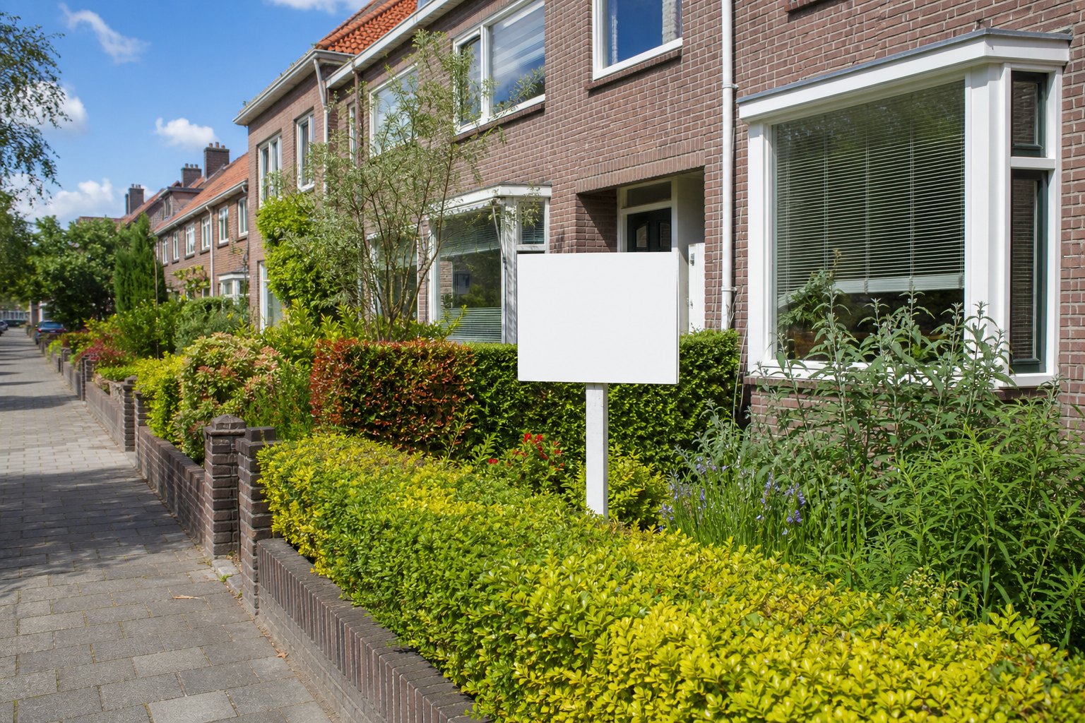 Nederlandse straat met bakstenen rijtjeshuizen en een leeg wit bordje in een voortuin